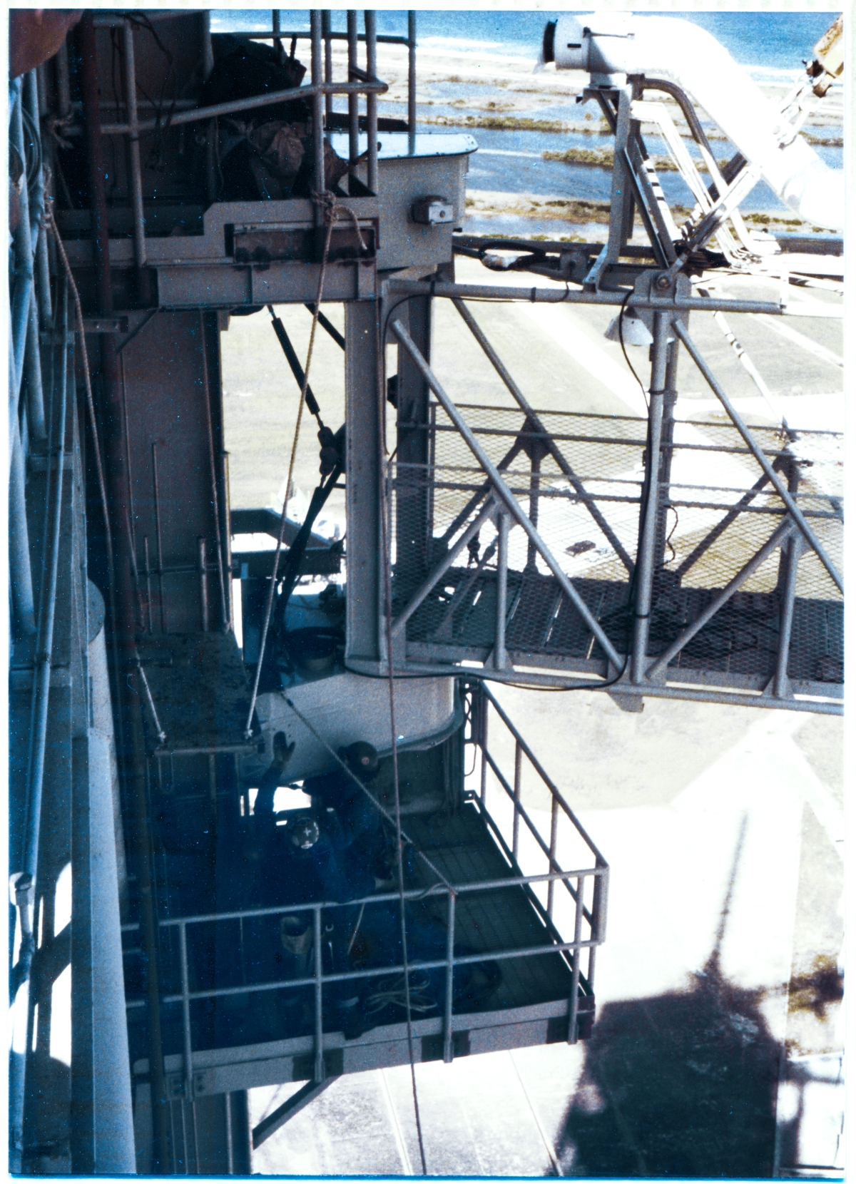 Image 114. At Space Shuttle Launch Complex 39-B, Kennedy Space Center, Florida, Union Ironworkers out of local Union Hall 808, working for Ivey Steel, have now gone metal-on-metal, attaching the GOX Arm to its Support Strongback near the top of the Fixed Service Structure. The near corner of the Lower Hinge Box Connection Plate has come in contact with the Strongback, and (poorly-visible in this old photograph) the right-hand ironworker on the Lower Hinge Access Platform has just shoved his sleever bar far enough through the matching bolt holes on the Arm and on the Strongback, to begin brute-force working the Arm into position, pinning it in place on the far bottom-right set of bolt holes. From here, the arm can be worked by the Crane Operator, the ironworker who is still on the ground working the tag line which is tied to the far end of the Arm, and other ironworkers on both the Upper and the Lower Hinge Access platforms using sleever bars, bull pins, or drift pins, to very gradually and very forcefully work it into its final orientation/position against the Strongback where all of the rest of the bolt holes in both the Connection Plates and on the Strongback will come into sufficient alignment to permit other ironworkers to start slipping bolts by hand, without damaging the threads on those bolts, through the aligned-holes in preparation for putting nuts on the ends of those bolts, at which point the nuts can begin to get torqued down. This will complete the job of finish-fastening the Arm to the tower, in its final location where it can be used to control and remove extremely-cold gaseous oxygen vapors which boil off from the liquid oxygen stored in the Tank, away from the area around the GOX Vent on the top of the Space Shuttle's External Tank, thereby preventing the build-up of ice formed from the high-humidity air at the Launch Pad in Florida, which could break off of the Tank during the violence of Lift-Off and fall, potentially impacting the delicate Thermal Protection System shielding the Orbiter, which is attached to the Tank below, from the metal-melting heat of re-entry, and possibly causing a catastrophic failure resulting in loss of vehicle and loss of crew. Photo by James MacLaren.